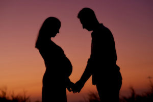 Couple expecting baby standing in sunset lights Side view silhouettes of unrecognizable pregnant woman and loving husband holding hands while standing face to face against colorful sundown sky in nature